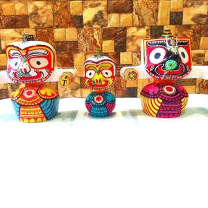 May include: Three colorful wooden figurines of deities with intricate details and painted faces. The figurines are arranged in a row on a white surface.