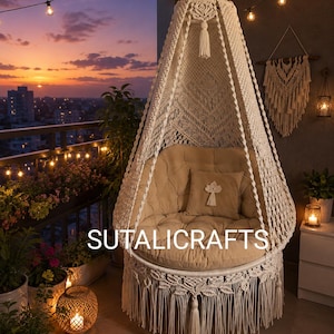 Peut inclure: Un fauteuil suspendu en macramé couleur crème avec un auvent conique, des motifs noués complexes et une longue frange. Le fauteuil comprend un coussin beige capitonné et un coussin carré assorti. Le texte SUTALICRAFTS est visible.