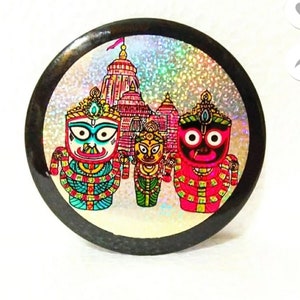 May include: A round, black-framed button with a colorful image of three Hindu deities in front of a temple. The deities are depicted in a traditional style with intricate details and bright colors. The background is a shimmering silver.