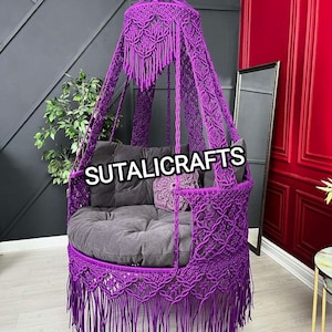 May include: Purple macrame hanging chair with a canopy and fringe. The chair is suspended from the ceiling and has a gray cushion.