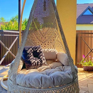 May include: A beige macrame hanging chair with a round cushion and a black pillow. The chair is hanging from a wooden frame and is outdoors on a patio.