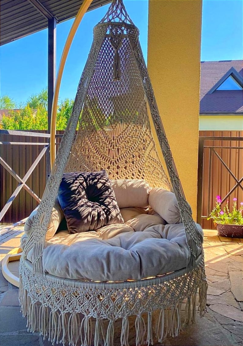 May include: A beige macrame hanging chair with a round cushion and a black pillow. The chair is hanging from a wooden frame and is outdoors on a patio.