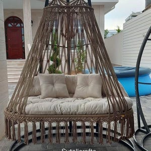 Op de afbeelding: Een bruine macramé schommelstoel met een kussen en twee vierkante kussens. De schommel hangt aan een zwart metalen frame en heeft decoratieve franjes. De tekst "Sutalicrafts" is zichtbaar aan de onderkant.
