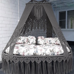 May include: A grey macrame hanging bed with a canopy. The bed has floral patterned cushions and pillows. The bed is suspended from chains and has a decorative fringe around the base. The canopy has a detailed woven design.