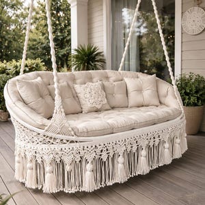 May include: A cream-colored macrame swing sofa with a curved back and a fringe trim. The sofa has several matching pillows and cushions. The swing is suspended by thick, white ropes and is designed for outdoor use. The sofa is on a wooden deck.