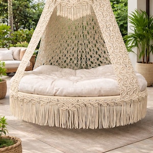 Peut inclure: Un daybed suspendu en macramé crème, avec une base et un auvent circulaires. Le lit est doté d'un coussin épais blanc cassé et est bordé de franges. L'auvent est soutenu par un motif en macramé tissé. Le daybed est à l'extérieur sur un patio.