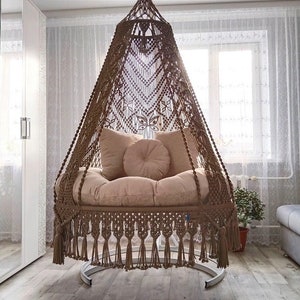 May include: A brown macrame hanging chair with a fringe border and a beige cushion. The chair is suspended from a white ceiling and is in a room with white walls and curtains.