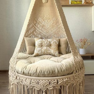 Op de afbeelding: Een beige macramé hangstoel met een ronde, gewatteerde zitting en rugleuning. De stoel heeft een decoratieve franje en is versierd met kussens in bijpassende tinten. Het ontwerp bevat ingewikkelde knoop patronen, waardoor een bohemien esthetiek ontstaat.