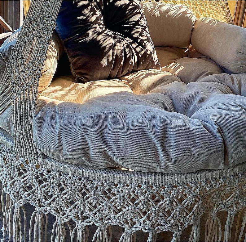 May include: A white macrame hanging chair with a gray cushion and a brown velvet pillow. The chair is made of thick rope and has a fringe border.