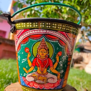 May include: A hand-painted metal bucket with a green handle. The bucket features a colorful depiction of a Hindu goddess sitting on a lotus flower. The goddess is wearing a red and yellow outfit and has four arms.