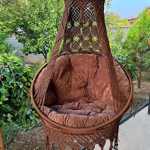 May include: A brown macrame hanging chair with a brown cushion. The chair is hanging from a ceiling hook and has a fringe around the bottom.