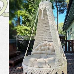 May include: A cream-colored macrame hanging chair with a round, tufted cushion. The chair has a canopy and decorative fringe. The chair is suspended from a wooden structure, with a natural outdoor background.