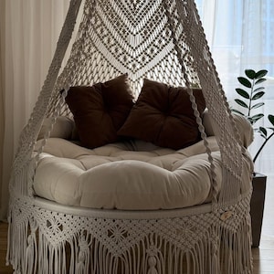 Op de afbeelding: Een crèmekleurige macramé hangstoel met een rond, gewatteerd kussen en twee bruine sierkussens. De stoel heeft een decoratieve franje langs de onderrand en is opgehangen aan een geweven luifel.