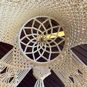 May include: A white macrame wall hanging with a circular design in the center. The macrame is made of thick cotton cord and has a fringe at the bottom.
