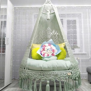 May include: A macrame hanging chair in a pale green colour, with a circular seat and a conical canopy. The chair is decorated with fringe and tassels. It has green and yellow cushions and a bouquet of pink and white flowers.