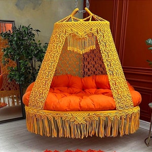 May include: A yellow macrame hanging chair with a bright orange cushion. The chair has a fringe border and is suspended from the ceiling.