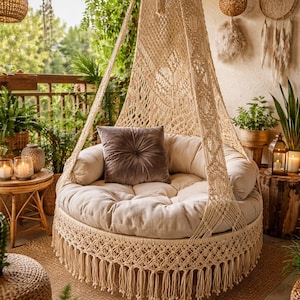 Peut inclure: Un daybed rond suspendu en macramé beige, avec une bordure à franges et un coussin moelleux. Un coussin en velours gris foncé repose sur le daybed. La scène extérieure comprend des plantes, des bougies et des guirlandes lumineuses.