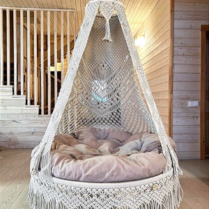 May include: A white macrame hanging chair with a fringe border and a thick, beige cushion. The chair is suspended from the ceiling in a room with wooden walls.