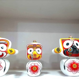 May include: Three white wooden figurines with painted faces and red, yellow, and black details. The figurines are standing on a white shelf.