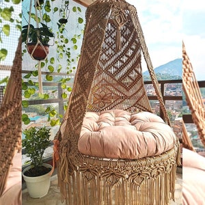 May include: A macrame hanging chair with a round, plush, light brown cushion. The chair is made of woven tan rope with a geometric pattern on the back and a fringe at the bottom. The chair is suspended on a balcony with plants.