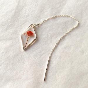 Puede incluir: Un pendiente de cadena de plata con un colgante en forma de diamante que presenta una flor roja prensada encapsulada en resina transparente.