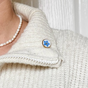 May include: A white sweater with a zip fastening and a small blue forget-me-not flower pin.