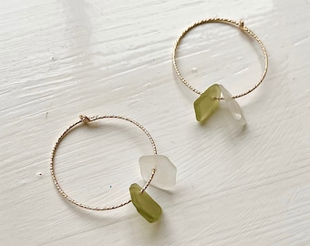 May include: A pair of gold hoop earrings with a twisted design. Each earring features two sea glass pieces, one light green and one clear, adding a touch of natural beauty. The earrings are displayed on a white surface.