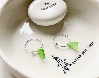May include: Two silver hoop earrings with green glass charms. The earrings are displayed in a white ceramic dish with the words "Be Yourself" and "Follow Your Heart" written on it.