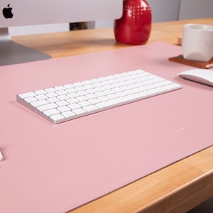 May include: A rectangular light pink desk mat with stitched edges, featuring a smooth surface. The mat is positioned on a wooden desk and holds a white wireless keyboard, a white computer mouse, a black smartphone, and white wireless earbuds.