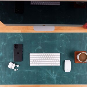 Op de afbeelding: Een teal lederen bureaumat met een wit toetsenbord, een witte muis, een zwarte smartphone, een witte draadloze oplader en witte oordopjes. Een bruine leren onderzetter met een witte koffiemok ligt op de mat.