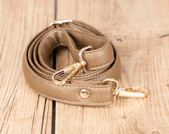 May include: A tan-colored purse strap with gold-colored metal clasps and hardware. The strap is made of a smooth, leather-like material and is stitched with white thread. The clasps are attached to the strap with a small, gold-colored button.