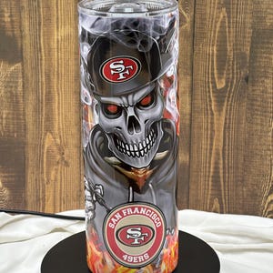 Może przedstawiać: Czarno-biała czaszka z czerwonymi oczami, nosząca czapkę w czarno-czerwonych barwach San Francisco 49ers. Czaszka otoczona jest płomieniami i dymem. Na dole kubka widnieje napis "San Francisco 49ers".