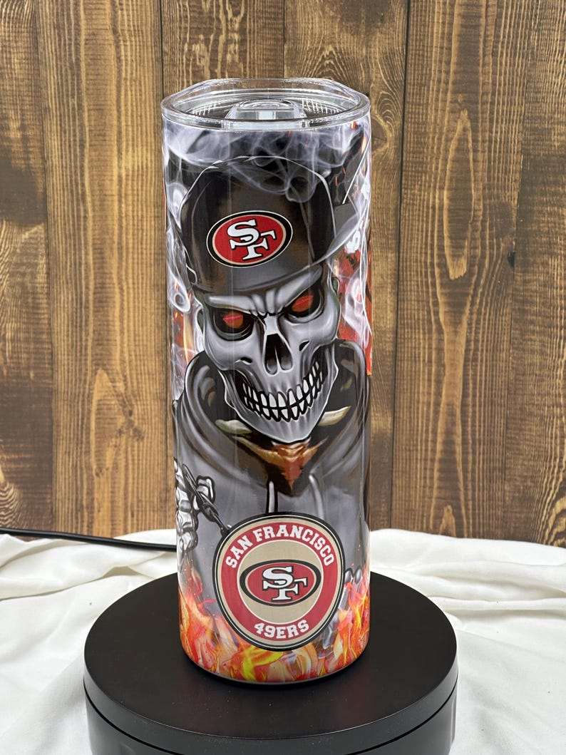 Może przedstawiać: Czarno-biała czaszka z czerwonymi oczami, nosząca czapkę w czarno-czerwonych barwach San Francisco 49ers. Czaszka otoczona jest płomieniami i dymem. Na dole kubka widnieje napis "San Francisco 49ers".