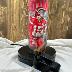 May include: A stainless steel tumbler with a red, white, and black design featuring a football player in a red jersey with the number 15. The tumbler has a black lid and a clear straw. The text "HOM" is visible on the tumbler.