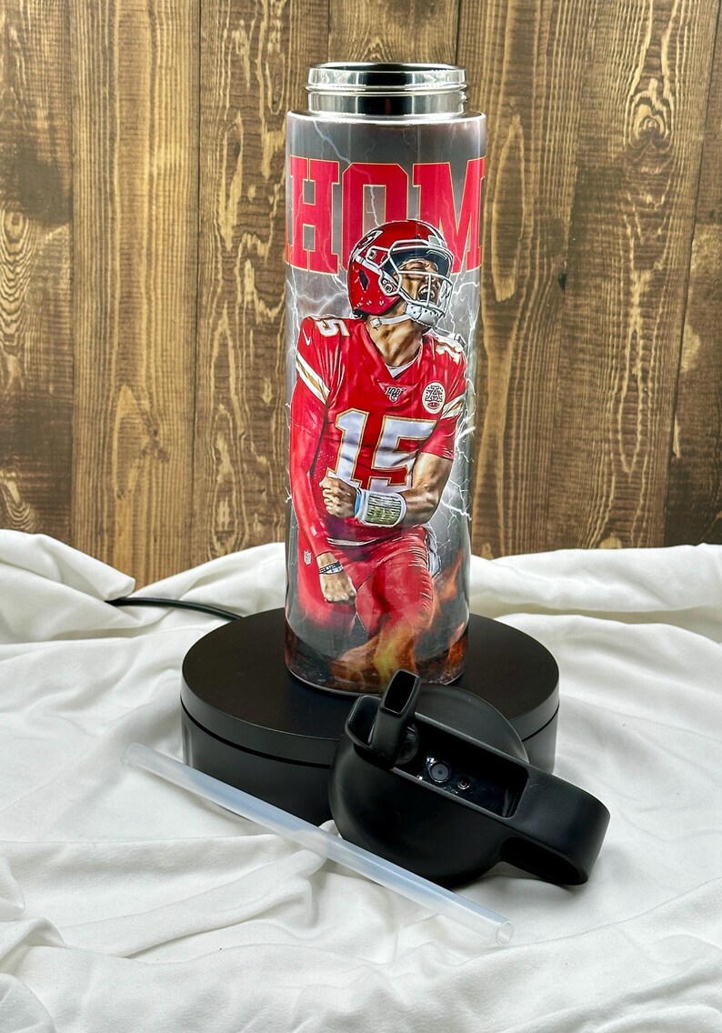 May include: A stainless steel tumbler with a red, white, and black design featuring a football player in a red jersey with the number 15. The tumbler has a black lid and a clear straw. The text "HOM" is visible on the tumbler.