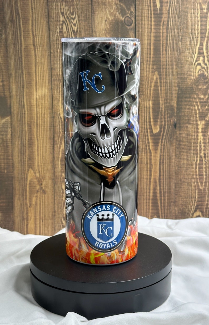 K&ouml;nnte beinhalten: Ein Edelstahlbecher mit einem Totenkopf-Design, der eine Baseballkappe mit den Buchstaben "KC" tr&auml;gt. Der Becher hat ein Kansas City Royals-Logo und einen feurigen Hintergrund. Der Becher ist etwa 20 cm hoch.