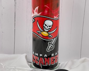 May include: A tall, cylindrical tumbler featuring the Tampa Bay Buccaneers logo. The design includes a skull with crossed swords and a football, set against a red and black background. The tumbler includes a lid, straw, and a black coaster.