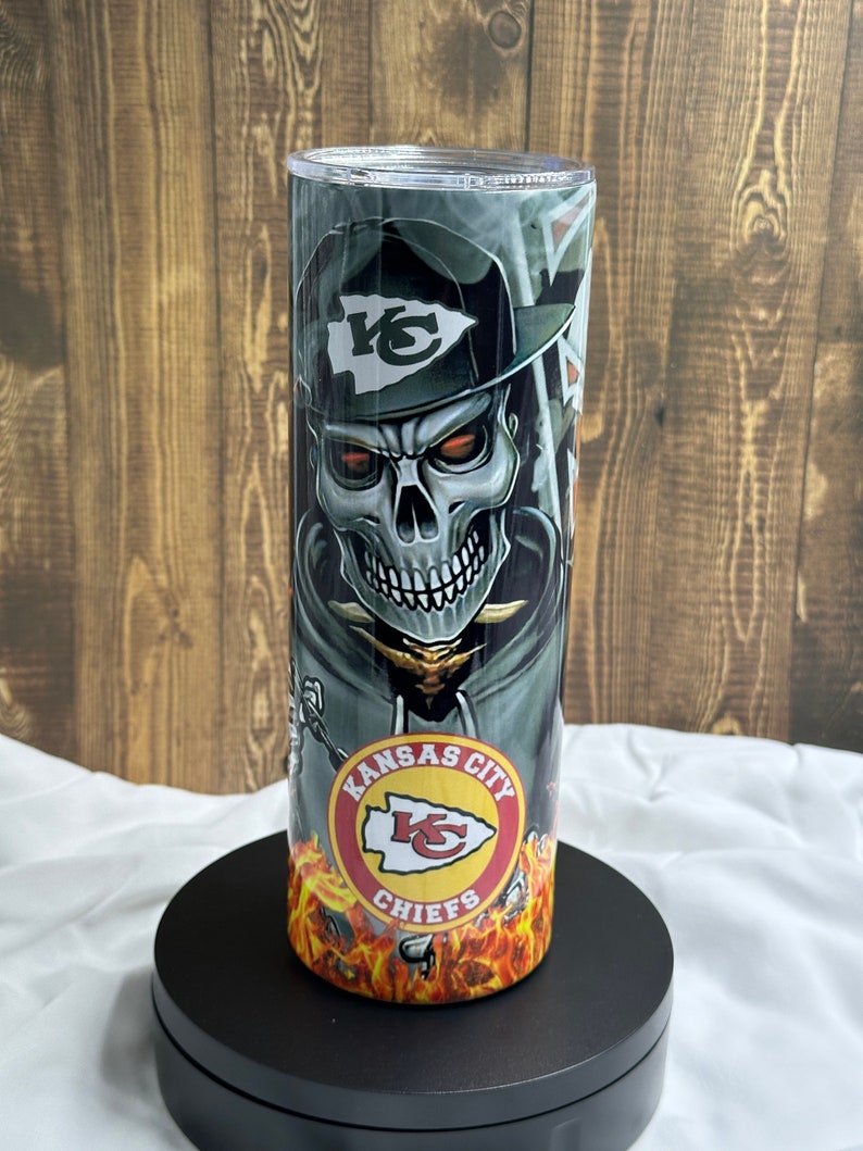 May include: A stainless steel tumbler featuring a skull wearing a Kansas City Chiefs hat and a red, white and yellow logo that reads "Kansas City Chiefs".