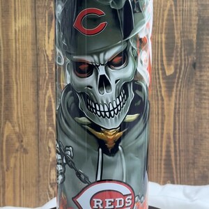 May include: A stainless steel tumbler with a skull wearing a baseball cap with the Cincinnati Reds logo. The skull is surrounded by flames and smoke. The tumbler has the words "REDS" printed on the bottom.