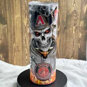 May include: A black and white tumbler with a skull wearing a baseball cap with the Arizona Diamondbacks logo. The skull is surrounded by flames. The tumbler is made of stainless steel and has a clear lid.