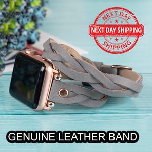 Puede incluir: Un reloj inteligente dorado con una correa de cuero trenzado gris. La correa se asegura con una hebilla dorada. El texto "GENUINE LEATHER BAND" se muestra en un cuadro negro.