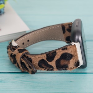 May include: A brown and black leopard print watch band with a silver buckle. The band is made of faux fur and is designed to fit a smartwatch.