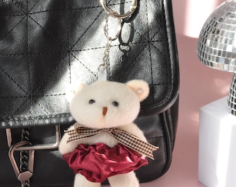 May include: A white teddy bear keychain with a burgundy skirt and a brown plaid bow is attached to a black quilted purse with a silver chain. The background is a soft pink.
