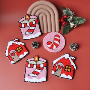 May include: Five Christmas-themed felt ornaments on a red background. The ornaments are a candy cane, a house, and three candles. The candles are pink with white and red stripes and a yellow flame.