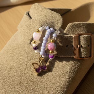 May include: A beige suede clog featuring a decorative charm accessory made of two linked beaded rings. The accessory includes light purple, white, and clear beads, gold-toned spacers, a heart-shaped bead, and a purple butterfly-shaped pendant.