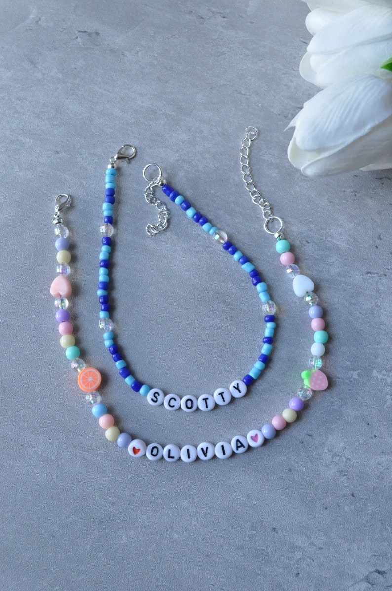 May include: Two beaded necklaces with colorful beads and charms. One necklace spells out "Olivia" with a heart charm and the other spells out "Scotty".