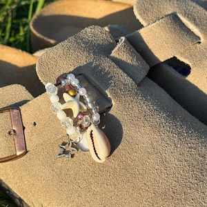 Può includere: Uno zoccolo in camoscio beige con un accessorio decorativo di perline fissato al cinturino. L'accessorio è composto da perline trasparenti, bianche e scure, una piccola stella marina bianca, un ciondolo a forma di stella argentata e una conchiglia.
