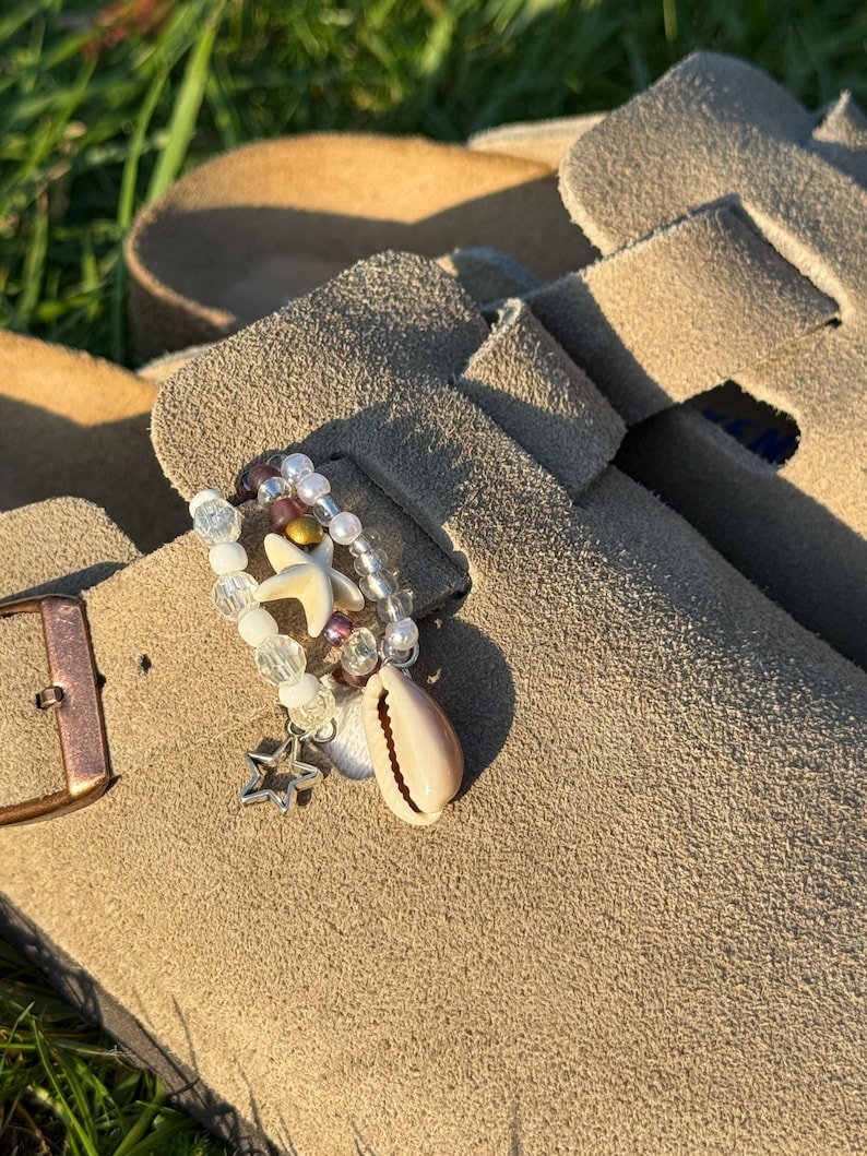 以下が含まれることがあります： ストラップにビーズの装飾アクセサリーが付いたベージュのスエードクロッグ。アクセサリーは、透明、白、ダークカラーのビーズ、小さな白いヒトデ、銀色の星のチャーム、クリーム色のタカラガイで構成されています。