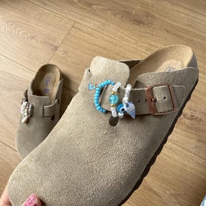 以下が含まれることがあります： コルクのフットベッドと銅色の金属製バックルが付いたトープ色のスエードクロッグ。各クロッグのストラップには、青いビーズ、白いパール、ハート、邪視除けのシンボルなど、小さな装飾チャームが複数取り付けられています。