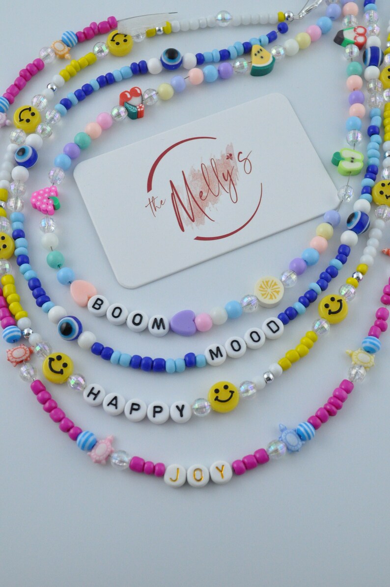 May include: Five colorful beaded necklaces with various charms, including smiley faces, hearts, fruit, and evil eyes. The necklaces spell out the words "BOOM", "MOOD", "HAPPY", and "JOY".